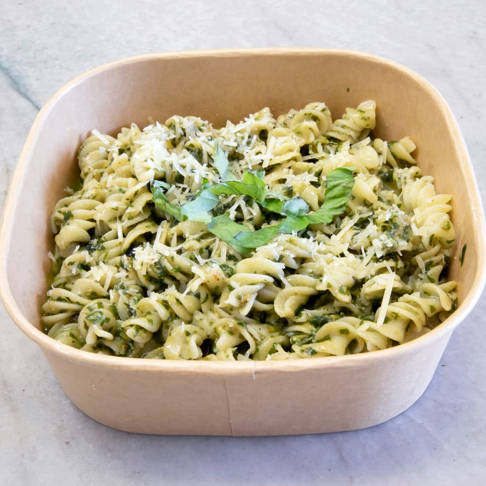 Pesto fusilli pasta with grated Parmesan cheese and fresh basil in a brown takeout container.