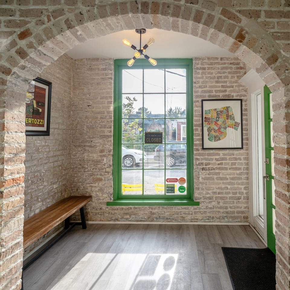 An arched brick entryway leads to a bright room with a large green-framed window and a wooden bench.