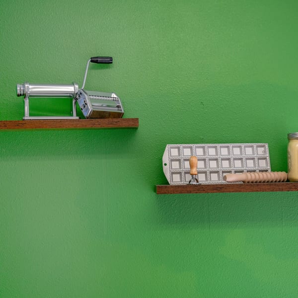 Pasta-making tools, including a pasta machine and ravioli mold, on wooden shelves against a green wall.