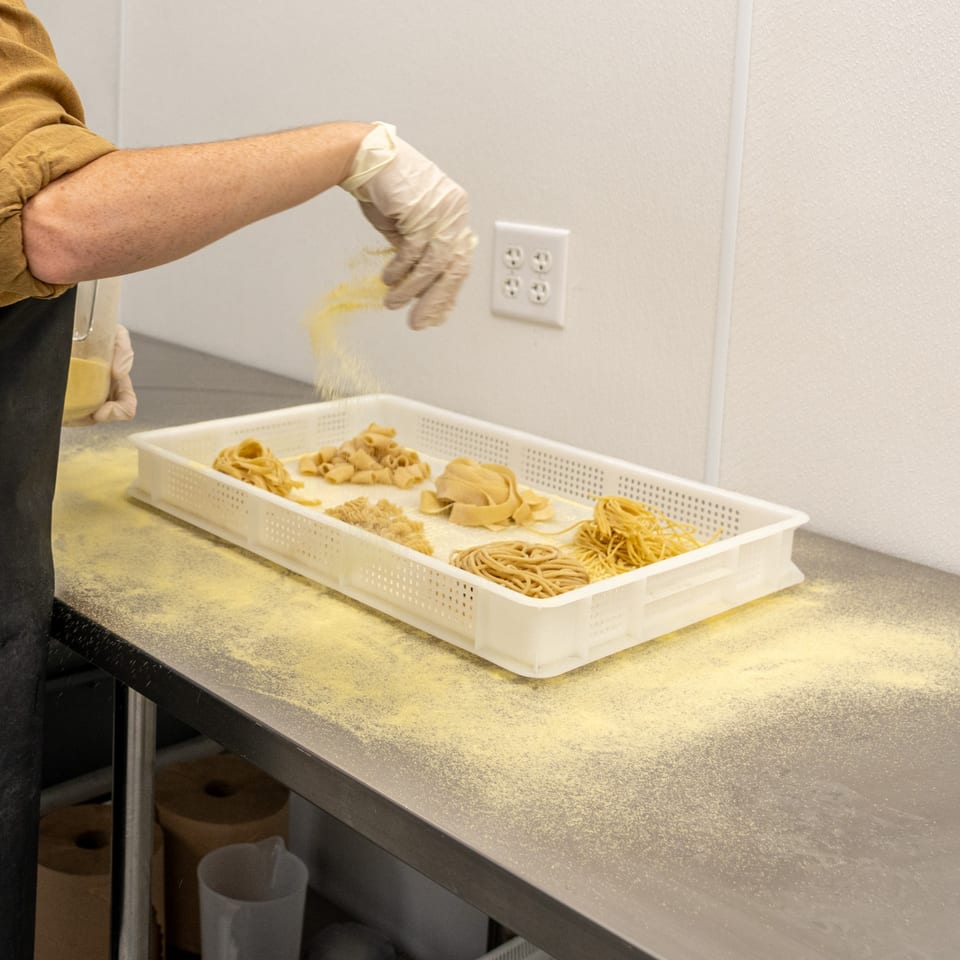 Gloved hands dusting fresh pasta varieties with flour in a white tray on a metal counter.