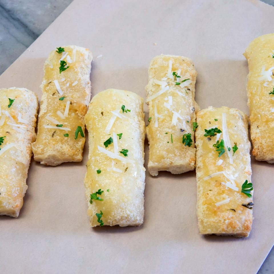 Golden-brown garlic breadsticks, topped with grated Parmesan cheese and fresh parsley, on a light brown surface.