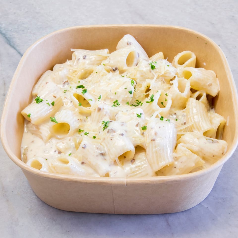 Creamy rigatoni pasta with grated cheese and parsley in a brown takeout container.
