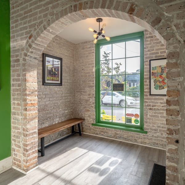 A brick archway leads into a bright room with a green-framed window and a wooden bench.