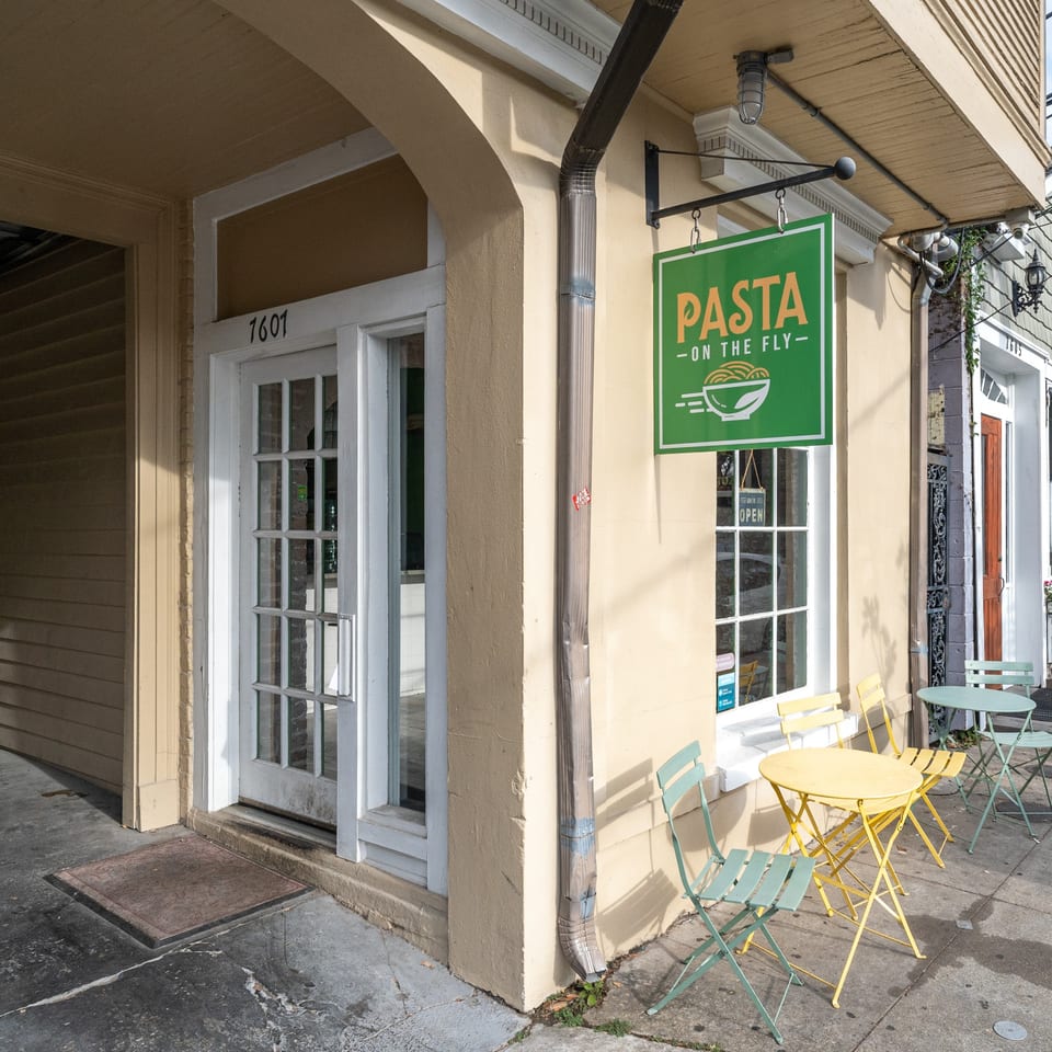 Exterior of Pasta On The Fly restaurant with green sign, yellow and green outdoor tables and chairs.