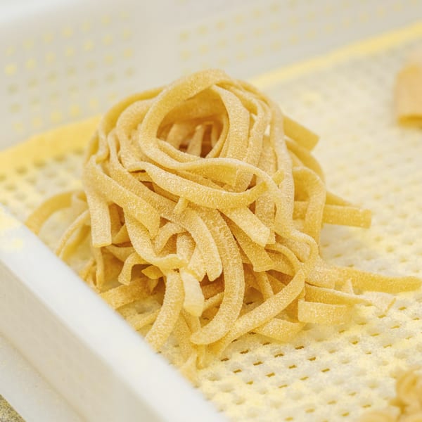 Freshly made fettuccine pasta, dusted with flour, rests on a textured white surface.