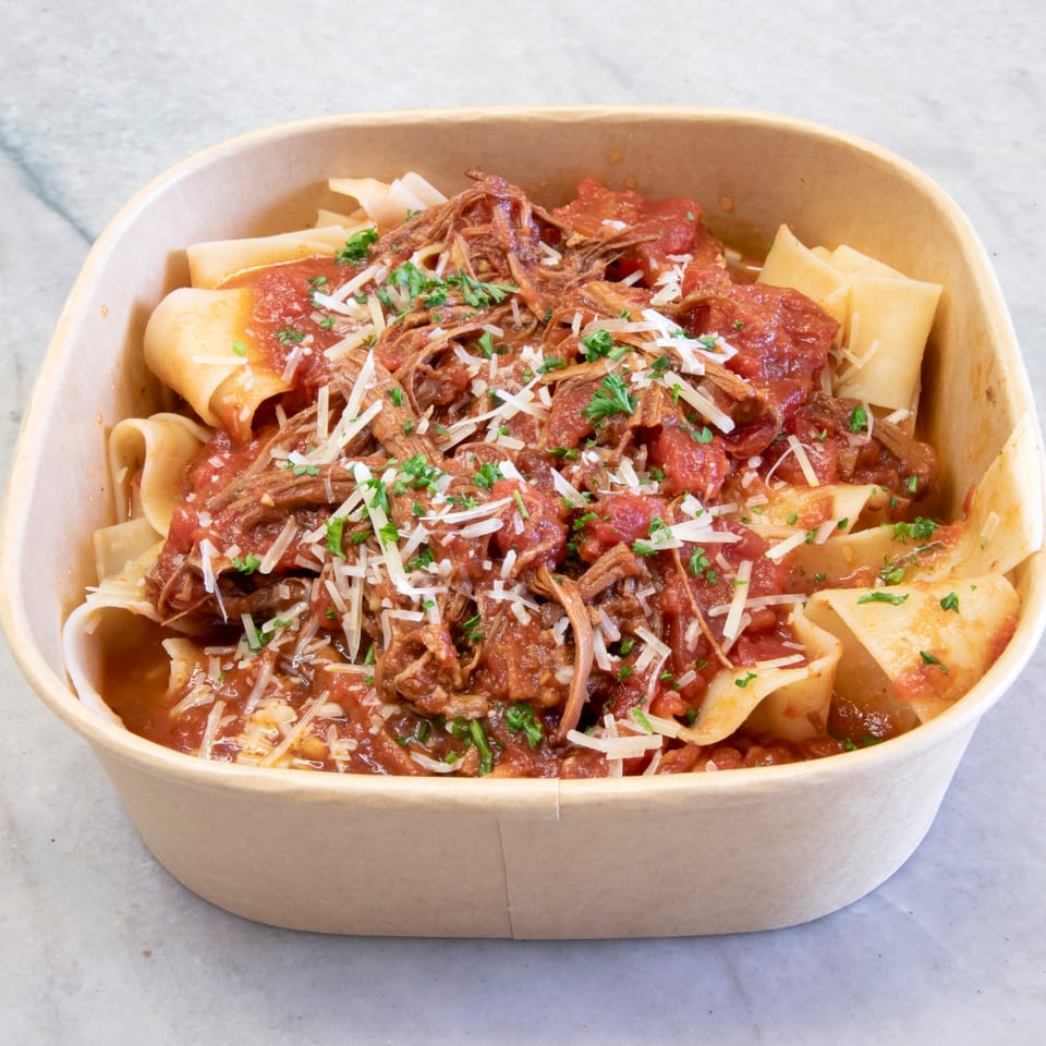 Pappardelle pasta with rich red sauce, shredded meat, parmesan, and parsley in a take-out container.