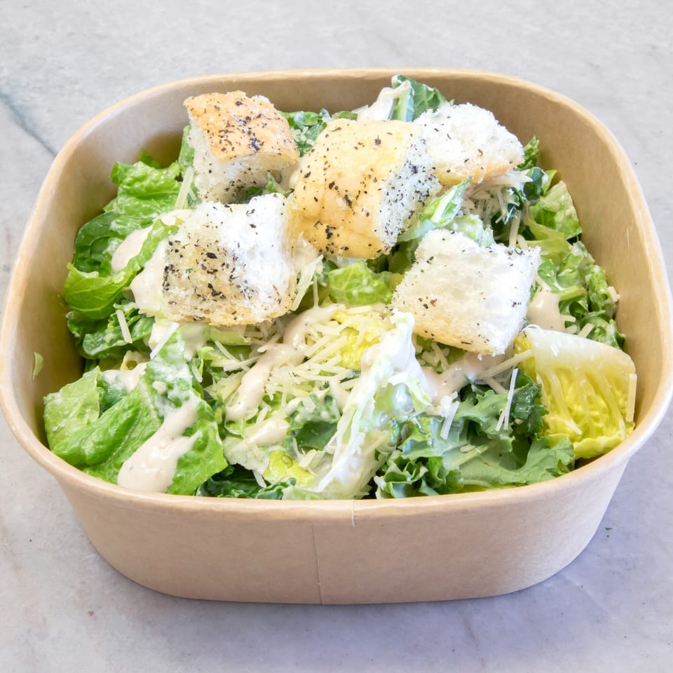 Caesar salad with romaine lettuce, croutons, parmesan cheese, and dressing in a brown takeout container.