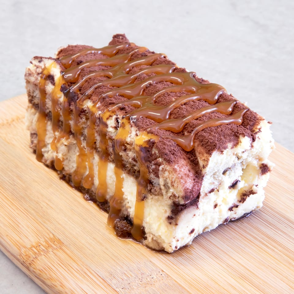A rectangular tiramisu dessert topped with cocoa powder and drizzled with caramel sauce on a wooden board.